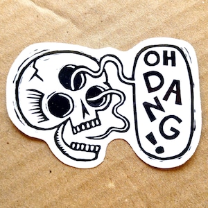 Op de afbeelding: Een zwart-witte sticker met een gestileerde schedel met de woorden "OH DANG!" in een tekstballon. De schedel heeft grote oogkassen en een gedetailleerde kaaklijn. De sticker ligt op een bruin oppervlak.
