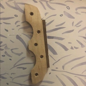 May include: A wooden guitar bridge with five holes drilled into it. The bridge is unfinished and has a natural wood color.