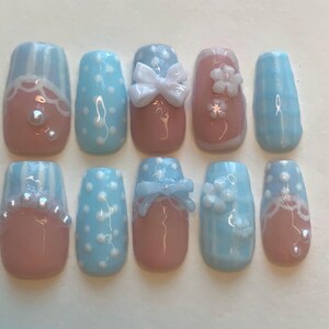 Puede incluir: Un conjunto de diez uñas artificiales con varios diseños. Las uñas presentan una combinación de colores azul claro, rosa y blanco. Algunas uñas tienen lunares, lazos o adornos florales. Otras tienen detalles de perlas o patrones de rayas.