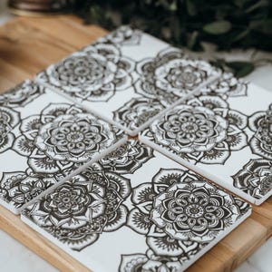 May include: Set of six square white ceramic coasters with a black floral mandala design. The coasters are arranged on a wooden cutting board. The coasters are a decorative and functional item for the home.
