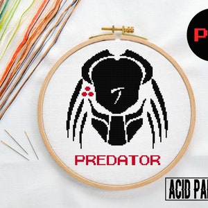Puede incluir: Un patrón de punto de cruz en blanco y negro del Predator de la franquicia de películas. El patrón está en un aro de bordado redondo con la palabra "PREDATOR" bordada debajo de la imagen. La imagen está sobre un fondo blanco con hilo de bordar y agujas visibles.