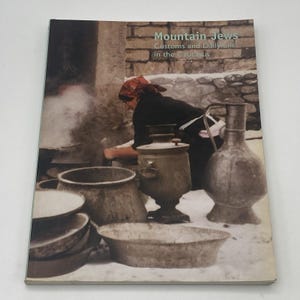 May include: A book titled "Mountain Jews Customs and Daily Life in the Caucasus" is displayed. The cover features a sepia-toned image of a person near cooking pots and a samovar. Various metal pots and a jug are also visible.