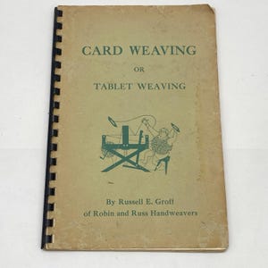 May include: A tan softcover book titled Card Weaving or Tablet Weaving by Russell E. Groff of Robin and Russ Handweavers. The cover features a green illustration of a person using a loom and is bound with a black plastic spiral spine.