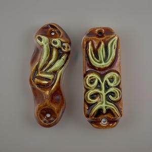 May include: Two brown ceramic wall plates with glossy finishes and light green embossed floral patterns. The left piece has an irregular, elongated shape, while the right piece is rectangular. Both feature small circular holes at the top and bottom.