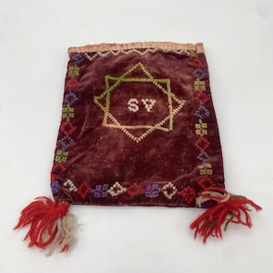May include: A small, burgundy velvet pouch with embroidered details. The pouch has a star-shaped design with the letters "SV" in the centre. The pouch has red and white tassels.