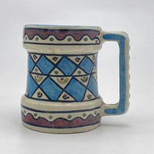 Könnte beinhalten: Ein Keramikbecher mit Henkel, mit cremefarbenem Grund und einem geometrischen Muster in Blau, Rot und Grün. Das Design des Bechers umfasst eine Reihe blauer Rauten und dekorativer Bänder.