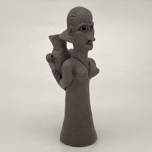 May include: A brown ceramic figurine of a woman carrying a large pot on her back. The woman has a stylized face with large eyes and a simple dress.