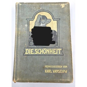 Puede incluir: Libro antiguo titulado "Die Schönheit" con una cubierta gris. La portada presenta una ilustración de una mujer y elementos florales. El texto "Die Schönheit" está en letras doradas, con el nombre del autor debajo.