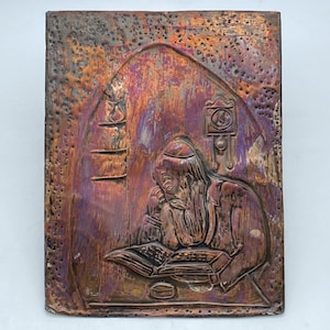 Könnte beinhalten: Rechteckige Kupferreliefskulptur, die eine Person beim Lesen eines Buches darstellt. Das Kunstwerk zeigt eine Figur mit langem Bart und Kopfbedeckung, die an einem Tisch sitzt. Die Farbpalette umfasst Kupfer-, Braun- und Lilatöne.