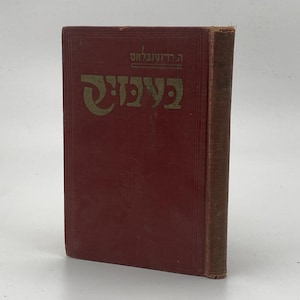 Pode incluir: Um livro de capa dura vintage com uma capa vermelho-escura apresentando texto hebraico em relevo dourado na frente. O título está escrito em letras douradas grandes e estilizadas, com texto hebraico menor acima e um emblema floral centralizado.