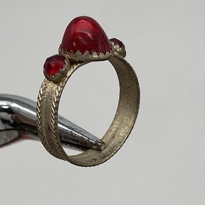 Puede incluir: Un anillo plateado con tres gemas rojas. La gema más grande es ovalada, y las otras dos son más pequeñas y redondas. La banda del anillo tiene un diseño texturizado. El anillo está sujeto por una herramienta plateada.