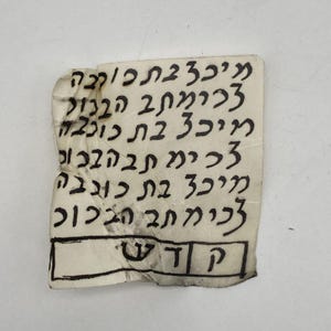 May include: A small, aged piece of paper with handwritten Hebrew text in black ink. The paper is torn and creased, with the text arranged in vertical columns. The bottom of the paper has a rectangular box with two Hebrew letters.