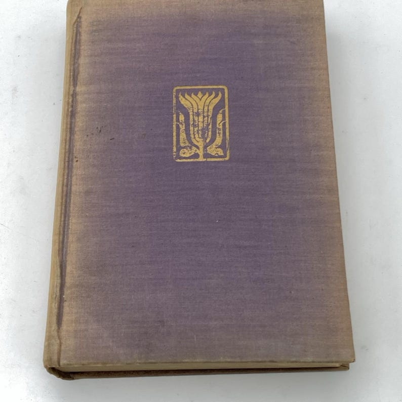May include: A vintage book with a purple cover and a gold emblem. The cover shows signs of wear, with a faded appearance. The gold emblem is a stylized floral design. The book's edges are slightly worn, suggesting age and use.
