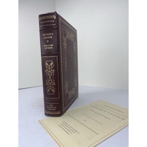 May include: A hardcover book titled "Sophie's Choice" by William Styron. The book is bound in dark burgundy leather with gold lettering and decorative accents. The words "First Edition" are at the top. A separate sheet of paper with the title is next to the book.