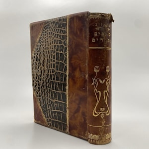 May include: A brown leather-bound book with a crocodile-embossed cover and gold lettering on the spine. The spine features a decorative design with flowers and leaves.