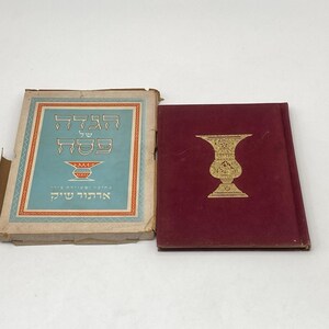 May include: A maroon hardcover book featuring a gold-stamped chalice on the front cover, accompanied by its original light blue and tan cardboard slipcase. The slipcase displays Hebrew text and a graphic illustration of a red and gold chalice.