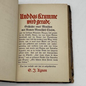 May include: Open book with text in German. The title "Und das Krumme wird gerade" is in red, with the author's name, S.J. Agnon, at the bottom. The pages are aged, with text in a serif font.
