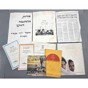 May include: A collection of printed materials, including books, pamphlets, and newspapers, with text in Hebrew and other languages. The items feature images of people and cultural scenes, with a yellow and red book in the foreground.