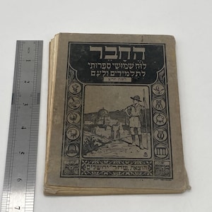 Puede incluir: Libro antiguo con texto hebreo e ilustraciones. La portada presenta una ilustración en blanco y negro de una persona y un paisaje urbano. El libro mide aproximadamente 15 cm de alto.