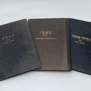 May include: Three rectangular books with dark covers arranged side-by-side. The left book is black, the center is brown, and the right is dark blue. Each cover features gold Hebrew text printed in the center, indicating titles and authorship.