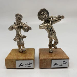 May include: Two silver figurines of men playing violins, each standing on a small rectangular base with a brown and beige marble-like surface. The bases have a label that reads "Ben-Zion Israel".