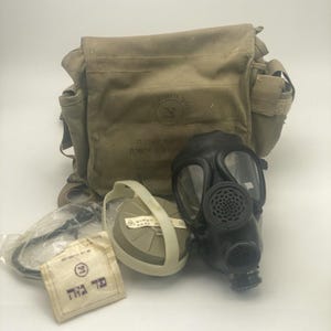May include: A military gas mask kit, comprising a black gas mask, a beige canvas bag, a filter, and additional components. The bag has a flap closure and pockets. The kit includes a filter and a small pouch with text.