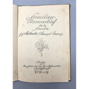 May include: An antique book, open to a page with ornate handwritten text and a floral illustration. The title "Formilion Shammbinh" is visible in elegant script. The pages are aged, and the cover is a dark green.