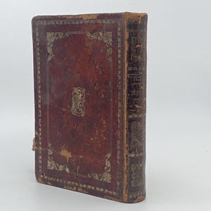 Puede incluir: Libro antiguo encuadernado en cuero, con una cubierta marrón rojiza desgastada. La cubierta presenta elementos decorativos dorados y un emblema central. El lomo muestra texto descolorido. Los bordes del libro muestran signos de edad y desgaste.