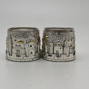 Puede incluir: Dos candelabros plateados con un relieve detallado de una escena urbana. El diseño incluye edificios con detalles dorados y un fondo texturizado. Los candelabros tienen un borde plateado y una base de piedra.