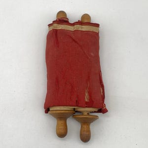 Könnte beinhalten: Eine kleine, antike Schriftrolle mit Holzgriffen und einer roten Stoffhülle. Der Stoff ist um die Holzdübel gewickelt, mit einem gold-cremefarbenen Band oben. Die Schriftrolle ist ein historisches Artefakt.