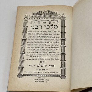 May include: An open book with Hebrew text, featuring ornate column-like designs and decorative elements. The page is aged, with visible text and a title that reads "Melchi Rabban". The book appears to be a religious text.