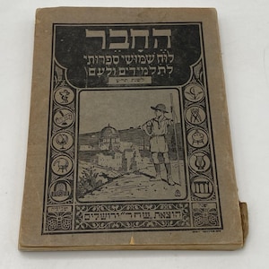 Puede incluir: Libro antiguo con texto e ilustraciones en hebreo. La portada presenta una pancarta negra con texto hebreo, un dibujo de una persona con un bastón y un paisaje urbano. El libro es de color marrón claro y muestra signos de desgaste.