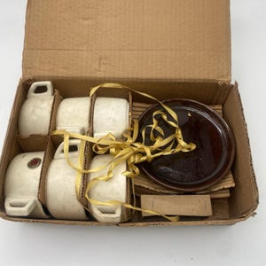 May include: A set of six white ceramic teacups and saucers with a brown saucer in a cardboard box. The cups and saucers are wrapped in yellow ribbon.