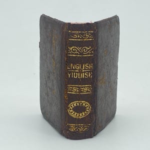 May include: A miniature antique book with a dark brown leather cover. The spine displays gold lettering: "ENGLISH YIDDISH", with decorative patterns. The book's edges show wear, indicating age. The overall appearance is vintage and classic, with a focus on the text.