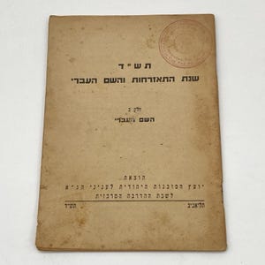 May include: A rectangular, tan-colored vintage booklet with Hebrew text printed in black. The cover features a circular stamp in the upper right corner and shows signs of age with light brown staining and worn edges throughout the paper surface.