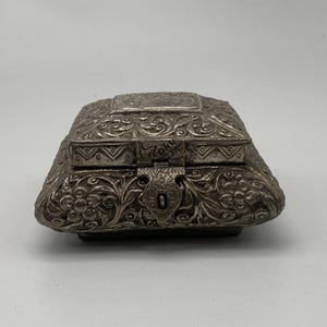 Peut inclure: Une boîte argentée ornée, avec un couvercle à charnière et un fermoir. La boîte présente des motifs floraux et de volutes détaillés sur sa surface. La boîte a une forme rectangulaire avec des bords arrondis et repose sur une petite base.