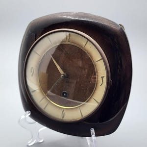 May include: A square-shaped desk clock with rounded corners, featuring a dark brown glossy frame. The clock face has a cream-colored outer ring with gold-toned hour markers and numerals at 12, 3, 6, and 9, surrounding a dark brown center.