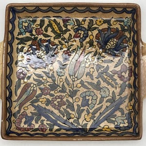May include: Square ceramic serving tray with a floral design. The tray has a beige background with a detailed pattern of flowers and leaves in shades of blue, red, and yellow. The rim is brown with a blue wavy border and handles.