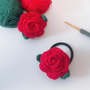 Puede incluir: Dos coleteros hechos a mano con rosas rojas de ganchillo y hojas verdes se muestran sobre una superficie blanca. Dos ovillos de lana, uno rojo y otro verde, están en el fondo. También se ve un ganchillo.