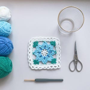 May include: A crocheted square with a blue floral pattern and white edging. Balls of blue and green yarn, a crochet hook, scissors, and a clear glass with a gold rim are arranged on a white surface. Crafting supplies are displayed.