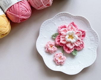 May include: A white scalloped plate displays handmade crochet flowers in shades of pink, white, and yellow, with green leaves. Several balls of yarn in pink, yellow, and green are in the background. The flowers are a craft project.