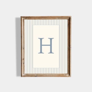 May include: A framed print with the letter "H" in a pale blue shade. The letter is centrally placed on a cream background, surrounded by a blue and white striped pattern. The frame is made of warm-toned natural wood.