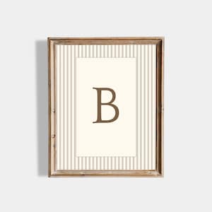 May include: A framed print with the letter "B" in a brown serif font. The artwork has a cream background with vertical brown pinstripes and a brown wooden frame. The design is classic and decorative, suitable for various interior styles.