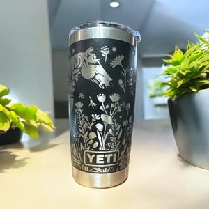 May include: A black stainless steel tumbler with a horse head and floral design etched on the side. The tumbler has a silver rim and the brand name "YETI" is displayed at the bottom. The tumbler is sitting on a light-coloured surface.