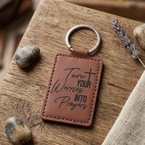 May include: A brown leather keyring with a silver ring. The rectangular tag features the text "Turn Your Worries Into Prayers" in a script font and a cross. The keyring is on a wooden surface with decorative stones.