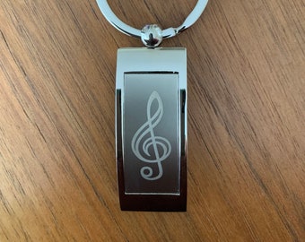 May include: A silver-colored keychain with a treble clef design. The rectangular pendant features an etched treble clef symbol. The keychain has a large, circular ring for keys.