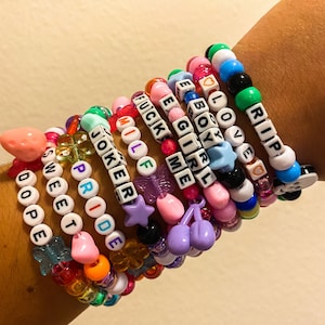 May include: A collection of colorful beaded bracelets with various charms and letter beads spelling out words like "DOPE", "SWEET", "PRIDE", "JOKER", "MILF", "FUCK ME", "E BOY", "GIRL", "LOVE", and "RIP". The bracelets feature a variety of colors and shapes.