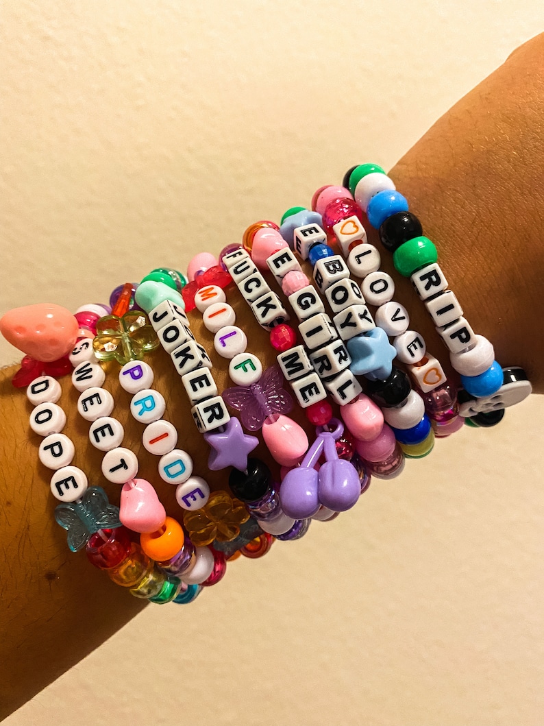 May include: A collection of colorful beaded bracelets with various charms and letter beads spelling out words like "DOPE", "SWEET", "PRIDE", "JOKER", "MILF", "FUCK ME", "E BOY", "GIRL", "LOVE", and "RIP". The bracelets feature a variety of colors and shapes.