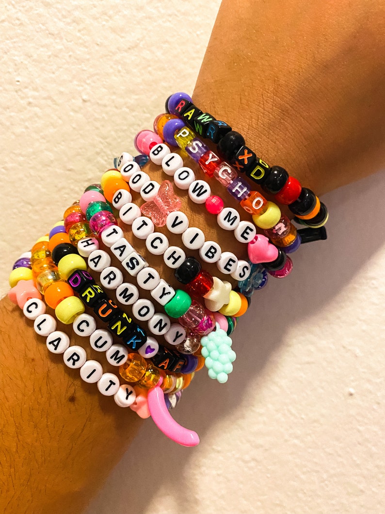 May include: A collection of colorful beaded bracelets stacked on a wrist. The bracelets feature various colors and include letter beads spelling out words like "RAW," "PSYCHO," and "CLARITY." Charms include a pink banana and a light blue bunch of grapes.