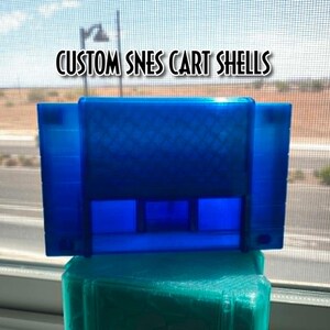 May include: A translucent blue plastic video game cartridge shell for a Super Nintendo Entertainment System. The text CUSTOM SNES CART SHELLS is printed above the cartridge. The shell features a rectangular shape with a textured grid pattern.